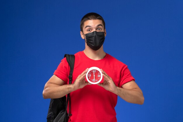 Front view male student in red t-shirt wearing backpack with mask holding clocks on blue background.