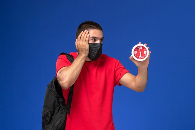 Front view male student in red t-shirt wearing backpack with mask holding clocks on blue desk.