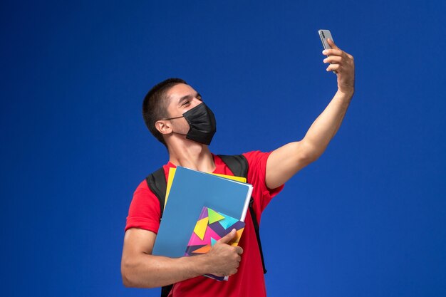 Front view male student in red t-shirt wearing backpack with mask holding files and taking selfie on blue background.