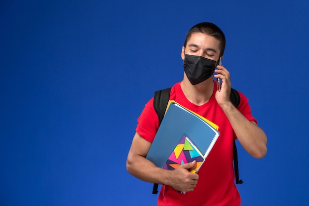 Front view male student in red t-shirt wearing backpack with mask holding files and talking on the phone on blue background.