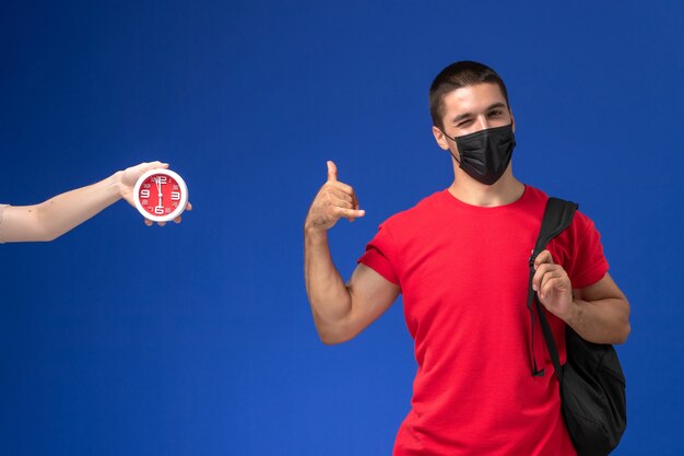 Front view male student in red t-shirt wearing backpack with mask posing on blue background.
