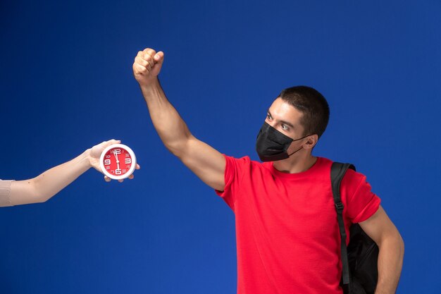 Front view male student in red t-shirt wearing backpack with mask posing on the blue background.