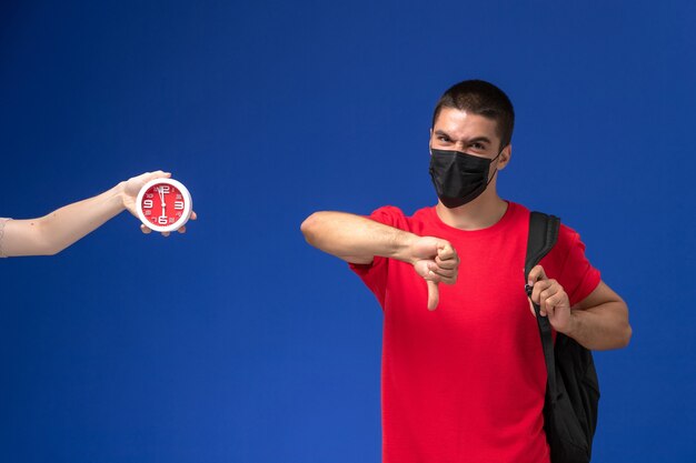 Студент вид спереди в красной футболке нося рюкзак с маской, позирующей на синем фоне.
