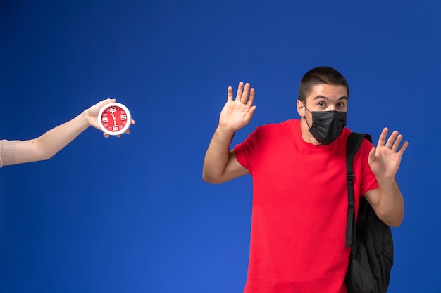 Front view male student in red t-shirt wearing backpack with mask scared of clocks on the blue background.