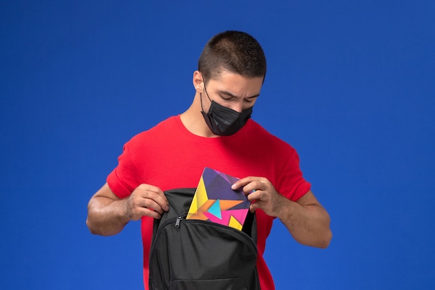 Front view male student in red t-shirt wearing mask and holding his backpack on the blue background.