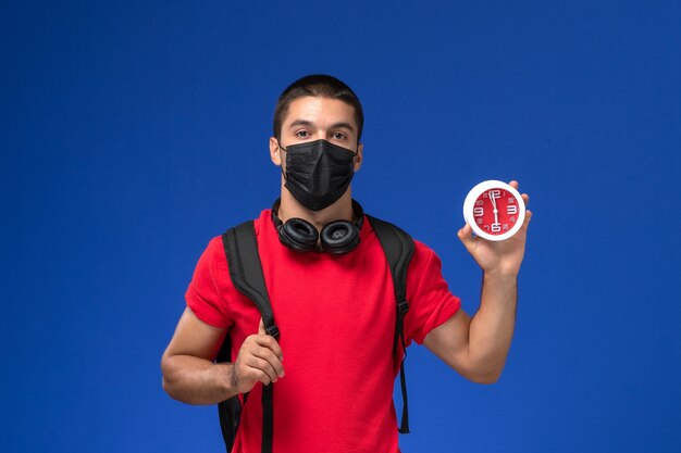 Front view male student in red t-shirt wearing mask with backpack holding clocks on blue background.