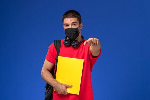青い背景に黄色のファイルを保持しているバックパックとマスクを身に着けている赤いTシャツの正面図男子学生。