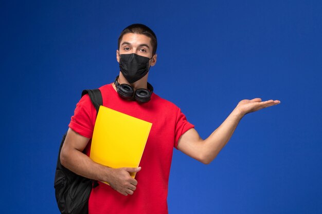 青い背景に黄色のファイルを保持しているバックパックとマスクを身に着けている赤いTシャツの正面図男子学生。