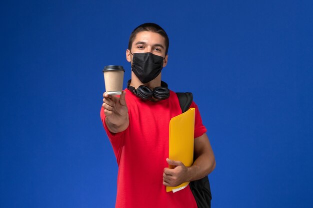 Front view male student in red t-shirt wearing mask with backpack holding yellow file and coffee on the blue background.