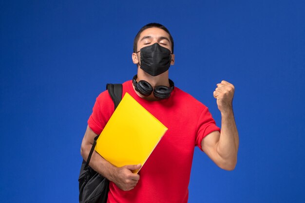 Студент вид спереди в маске красной футболки нося с рюкзаком, держащим ликование желтого файла на синем фоне.