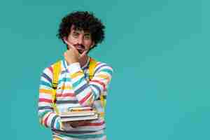 Free photo front view of male student in striped shirt wearing yellow backpack holding books and thinking on the blue wall