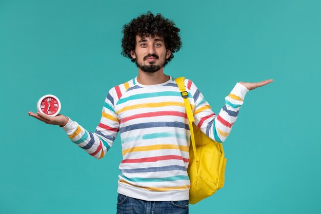 Front view of male student in striped shirt wearing yellow backpack holding clocks on blue wall