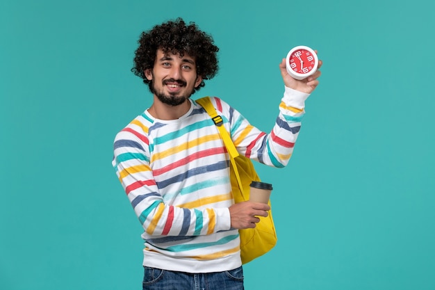 Foto gratuita vista frontale dello studente maschio in camicia a righe che porta zaino giallo che tiene caffè e orologi sulla parete blu