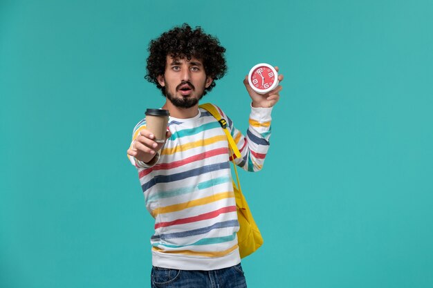 Front view of male student in striped shirt wearing yellow backpack holding coffee and clocks on the blue wall