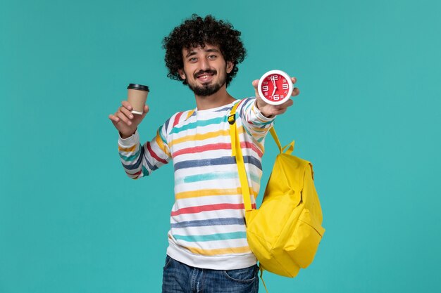 Front view of male student in striped shirt wearing yellow backpack holding coffee and clocks smiling on blue wall