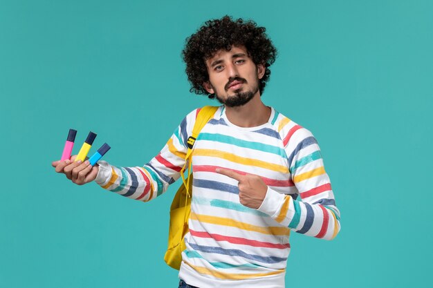 Front view of male student in striped shirt wearing yellow backpack holding colored felt pens on blue wall