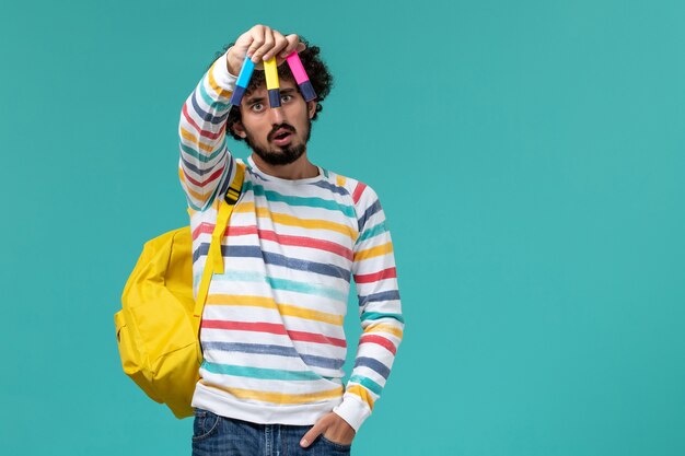 Front view of male student in striped shirt wearing yellow backpack holding colored felt pens on the blue wall