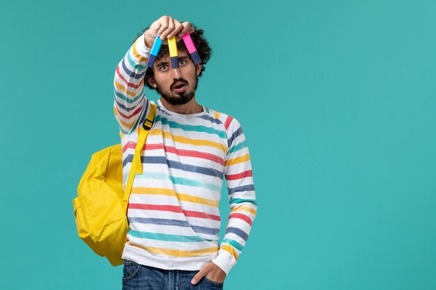 Студент в полосатой рубашке, одетый в желтый рюкзак, держит цветные фломастеры на синей стене, вид спереди