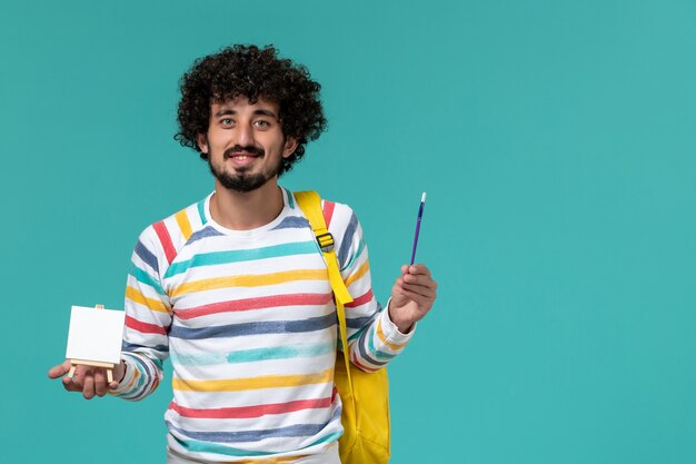 Front view of male student in striped shirt wearing yellow backpack holding easel and tassel on blue wall