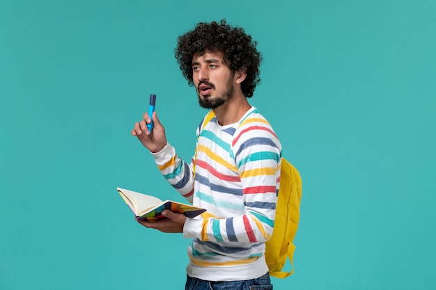 青い壁にフェルトペンとコピーブックを保持している黄色のバックパックを身に着けている縞模様のシャツの男子学生の正面図