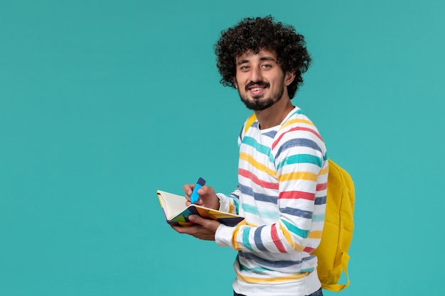 Foto gratuita vista frontale dello studente maschio in camicia a righe che porta zaino giallo che tiene pennarello e quaderno sulla parete blu