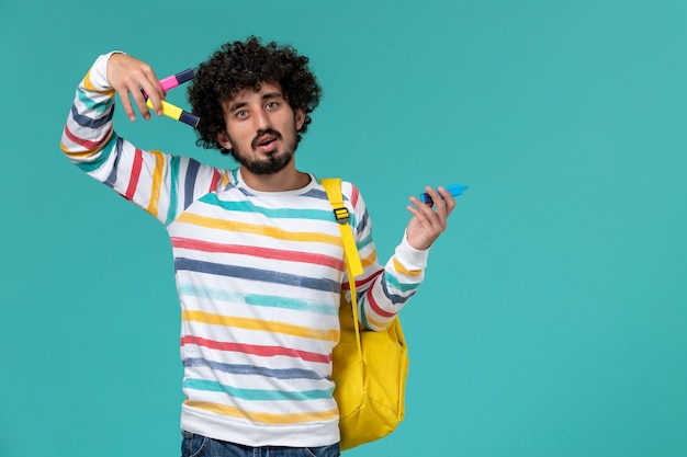 Front view of male student in striped shirt wearing yellow backpack holding felt pens on blue wall