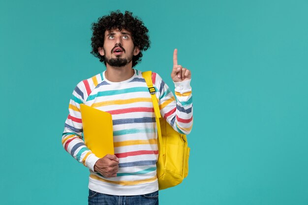 Front view of male student in striped shirt wearing yellow backpack holding files on blue wall