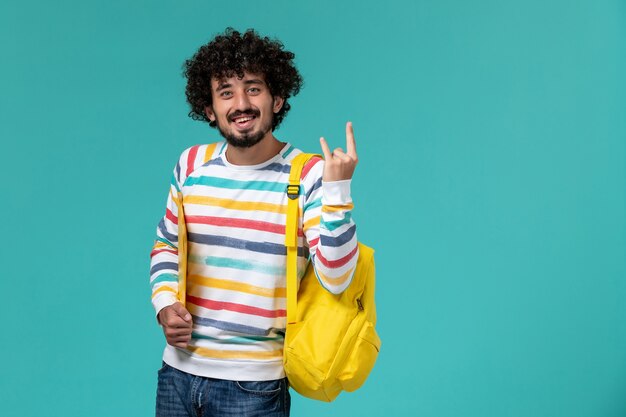 Front view of male student in striped shirt wearing yellow backpack holding files on the blue wall