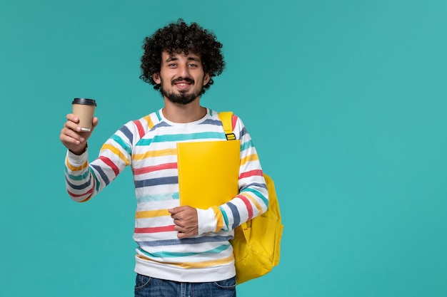 Front view of male student in striped shirt wearing yellow backpack holding files and coffee on blue wall