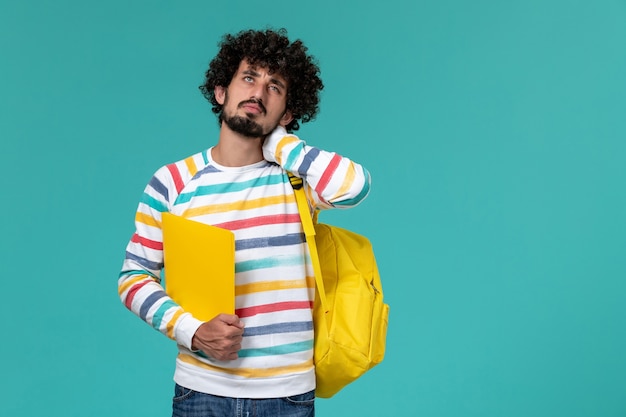 Vista frontale dello studente maschio in camicia a righe che indossa lo zaino giallo che tiene i file con mal di testa sulla parete blu