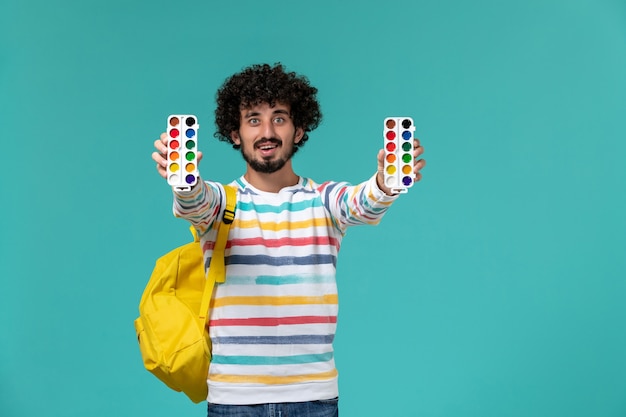 Front view of male student in striped shirt wearing yellow backpack holding paints on blue wall