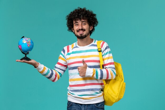 Front view of male student in striped shirt wearing yellow backpack holding round little globe on blue wall