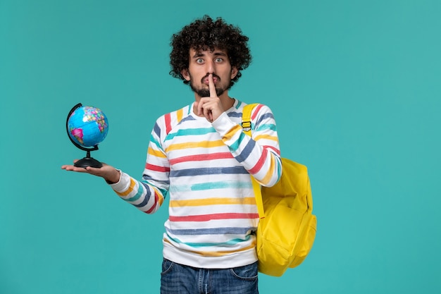 Foto gratuita vista frontale dello studente maschio in camicia a strisce che porta zaino giallo che tiene piccolo globo rotondo sulla parete blu