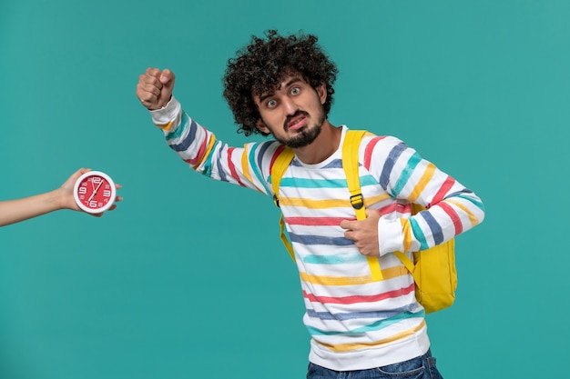 Foto gratuita vista frontale di uno studente maschio in camicia a righe che indossa uno zaino giallo in posa sulla parete blu chiaro