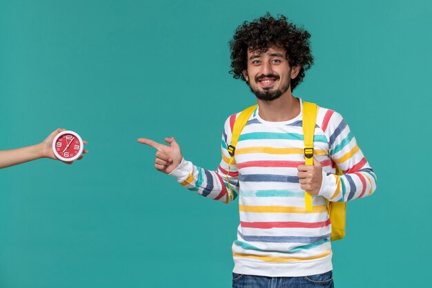 Front view of male student in striped shirt wearing yellow backpack posing and smiling on light blue wall