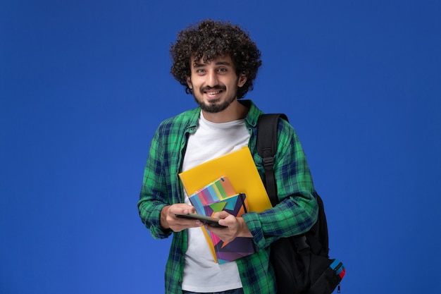 Вид спереди студента в черном рюкзаке, держащего тетрадь и файлы, использующего свой телефон на голубой стене