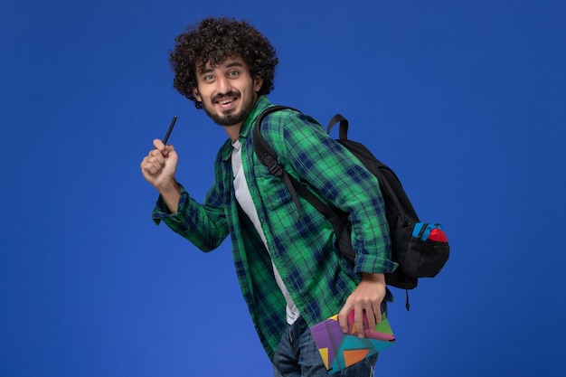 Front view of male student wearing black backpack holding copybook and pen running on blue wall