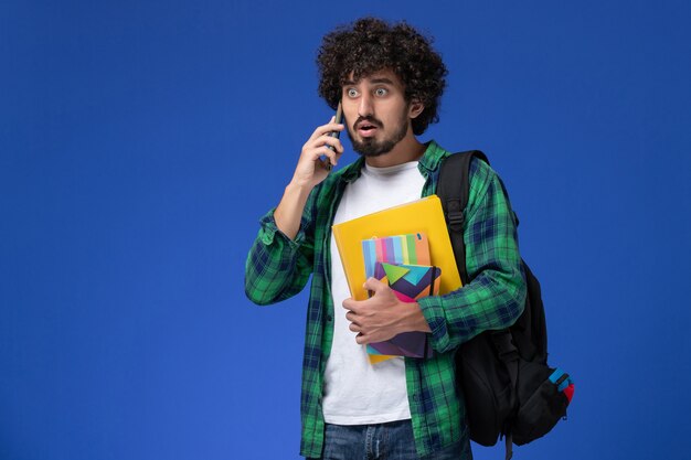 水色の壁に電話で話しているコピーブックとファイルを保持している黒いバックパックを身に着けている男子学生の正面図