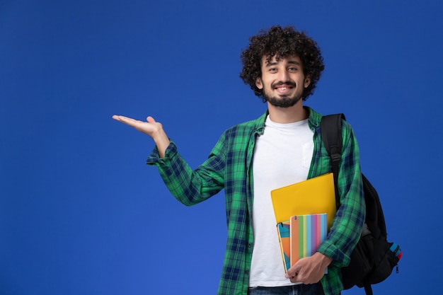 Вид спереди студента в черном рюкзаке, держащего тетради и улыбающегося на синей стене