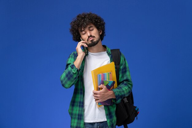 Vista frontale dello studente maschio che indossa uno zaino nero che tiene file e quaderni parlando al telefono sulla parete azzurra