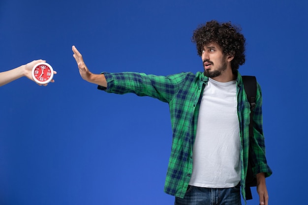 Front view of male student wearing black backpack posing with confused expression on the light blue wall