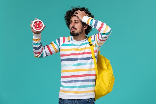 Front view of male student wearing yellow backpack holding clock on the light blue wall