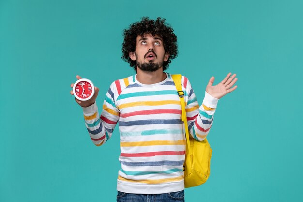 Front view of male student wearing yellow backpack holding clocks on light blue wall