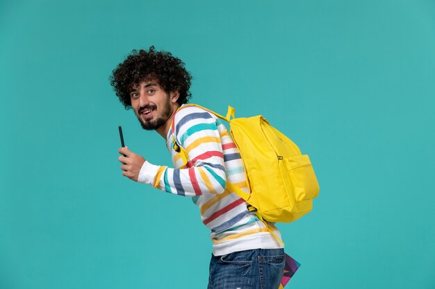 青い壁にコピーブックとペンを保持している黄色のバックパックを身に着けている男子学生の正面図