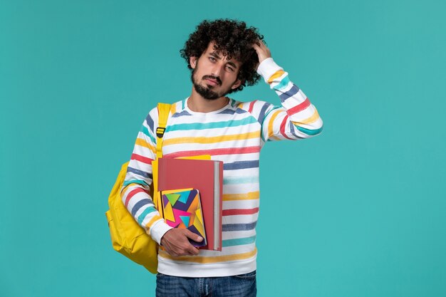 Front view of male student wearing yellow backpack holding files and copybook on blue wall