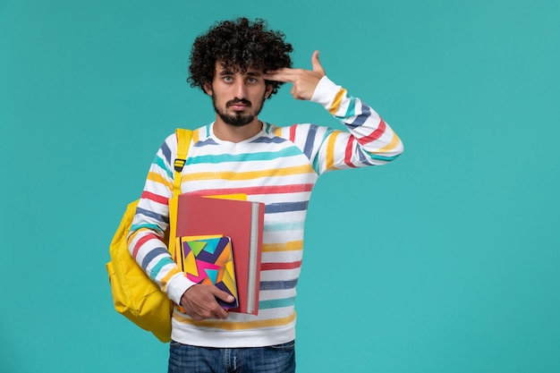 Front view of male student wearing yellow backpack holding files and copybook on blue wall