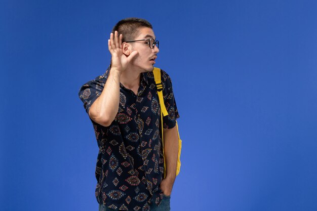 Вид спереди студента в желтом рюкзаке, пытающегося услышать на синей стене