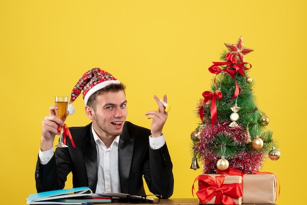 Вид спереди мужчина-работник празднует рождество с шампанским вокруг маленькой рождественской елки и подарками на желтом столе новогодний офисный цвет рождество