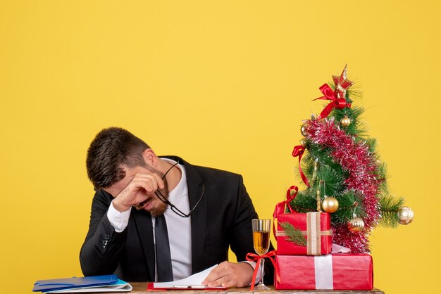 Front view male worker behind his table with presents and xmas tree stressed on yellow 