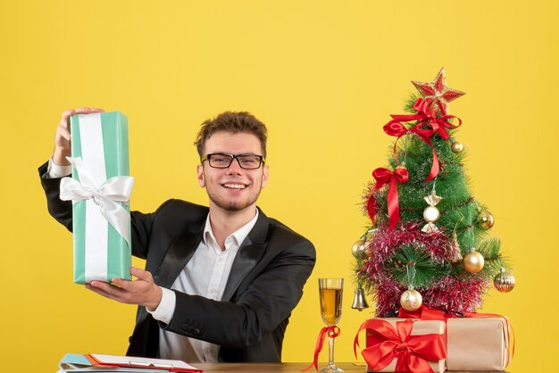 Front view male worker behind his working place holding present in blue package on yellow 